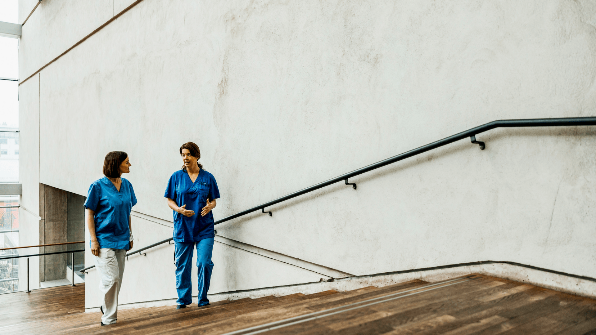 Deux professionnelles de la santé en tenue médicale bleue marchent ensemble et discutent dans un escalier en bois d’un corridor d’hôpital moderne et lumineux. Deux professionnelles de la santé en tenue médicale bleue marchent ensemble et discutent dans un escalier en bois d’un corridor d’hôpital moderne et lumineux.