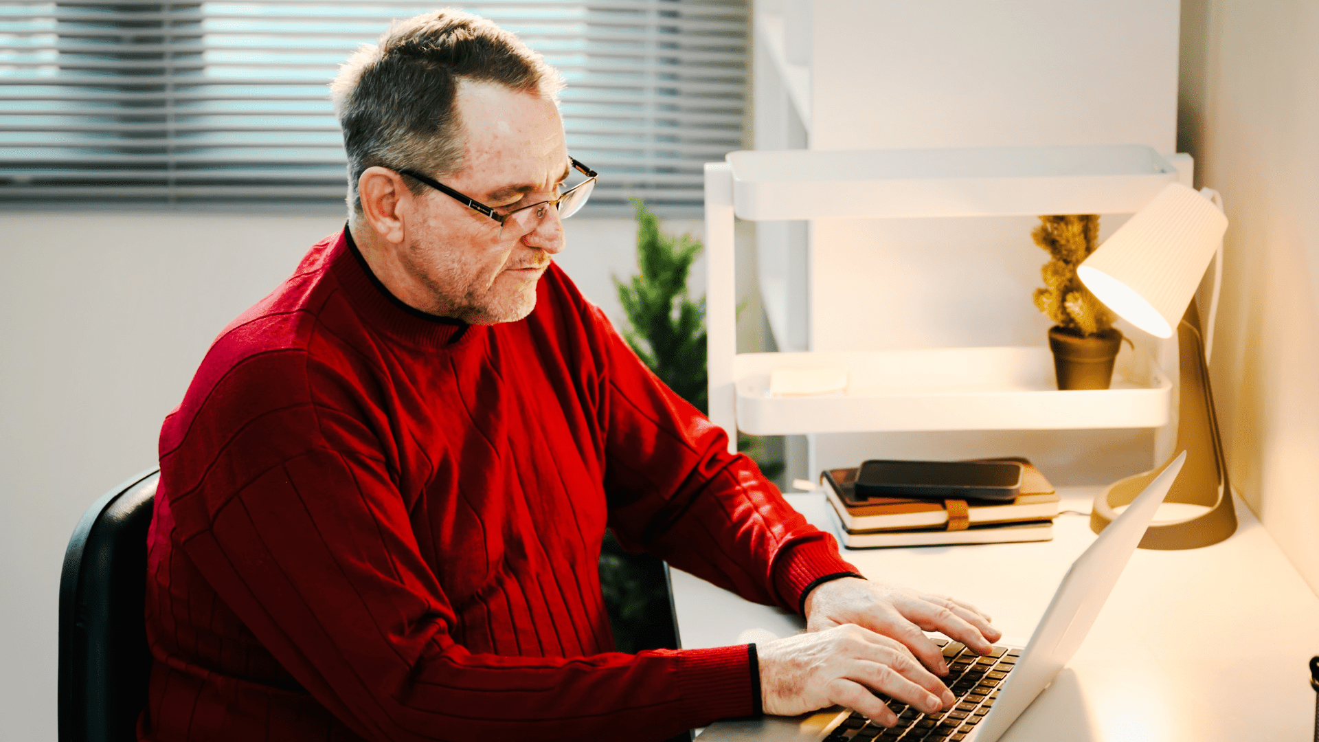 Man in red sweater working on a laptop at minimalist desk under desk lamp