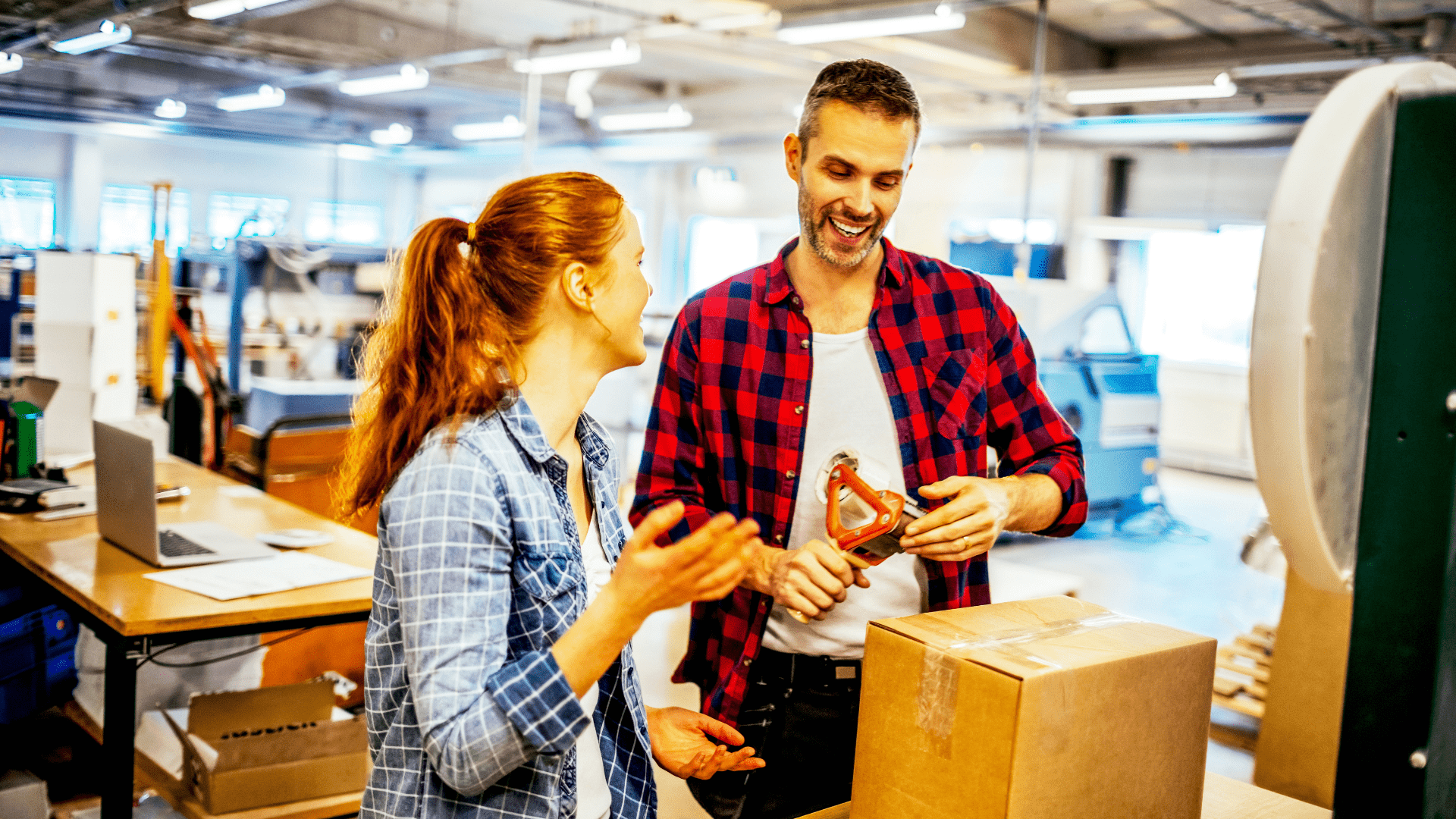 Two coworkers smiling and talking in a workshop, with the man holding a handheld power tool beside a large cardboard box.