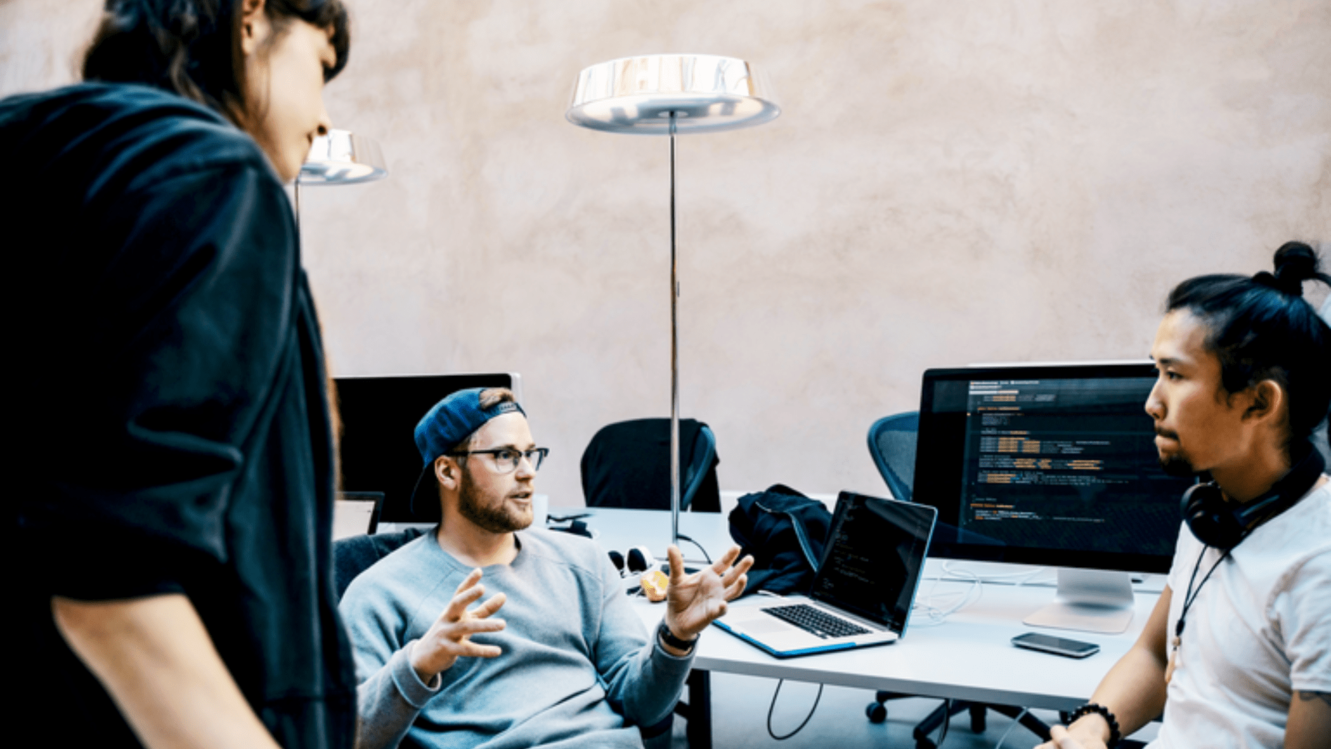 individuals discussing at a work table, next to computer monitors