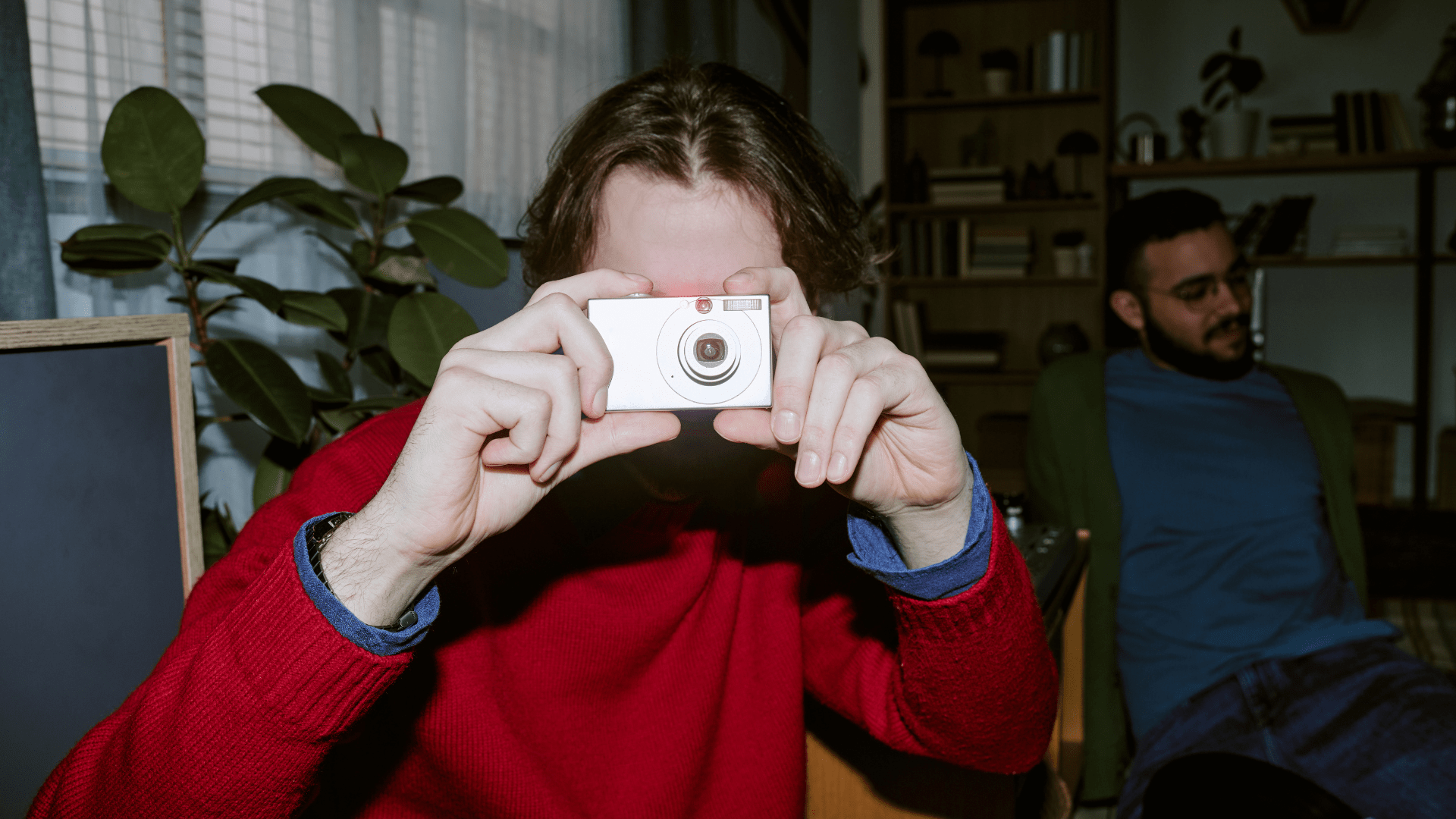 Une personne portant un chandail rouge tient un petit appareil photo devant son visage, tandis qu’une autre personne est assise à l’arrière-plan près d’une bibliothèque et de plantes d’intérieur.