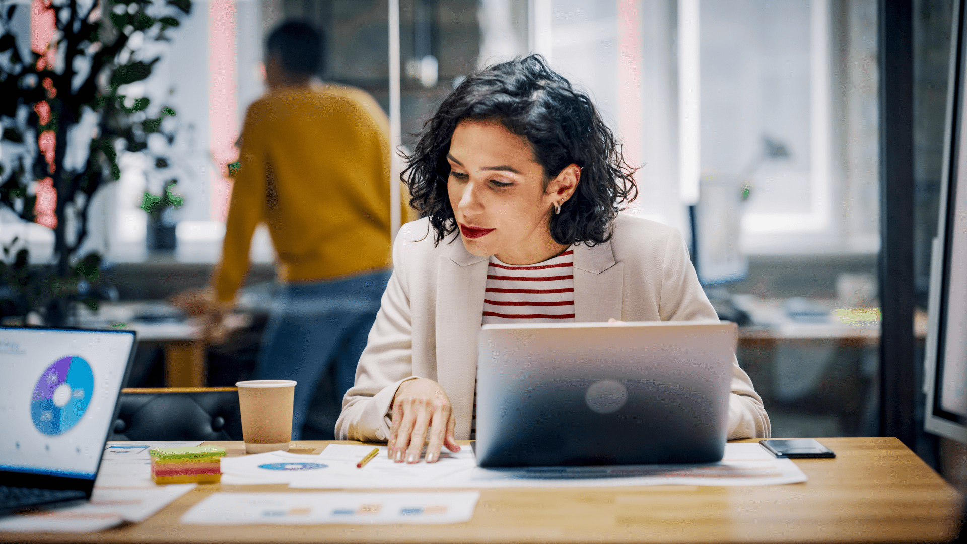 Une femme aux cheveux courts et foncés est assise à son bureau près d’un ordinateur portable ouvert, examinant des graphiques imprimés disposés sur la table.