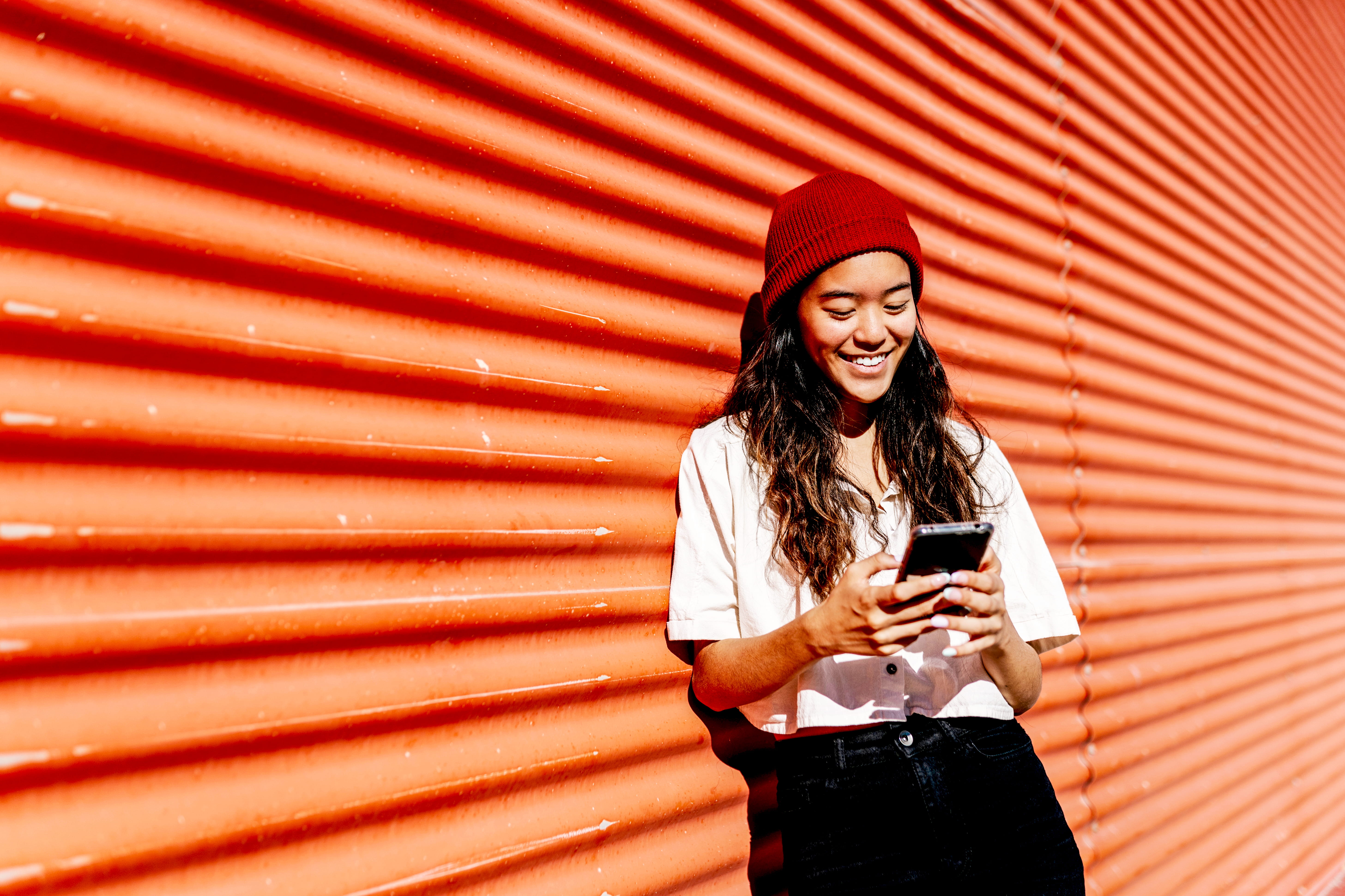 Femme souriante utilisant son téléphone mobile devant un mur rouge en tôle ondulée