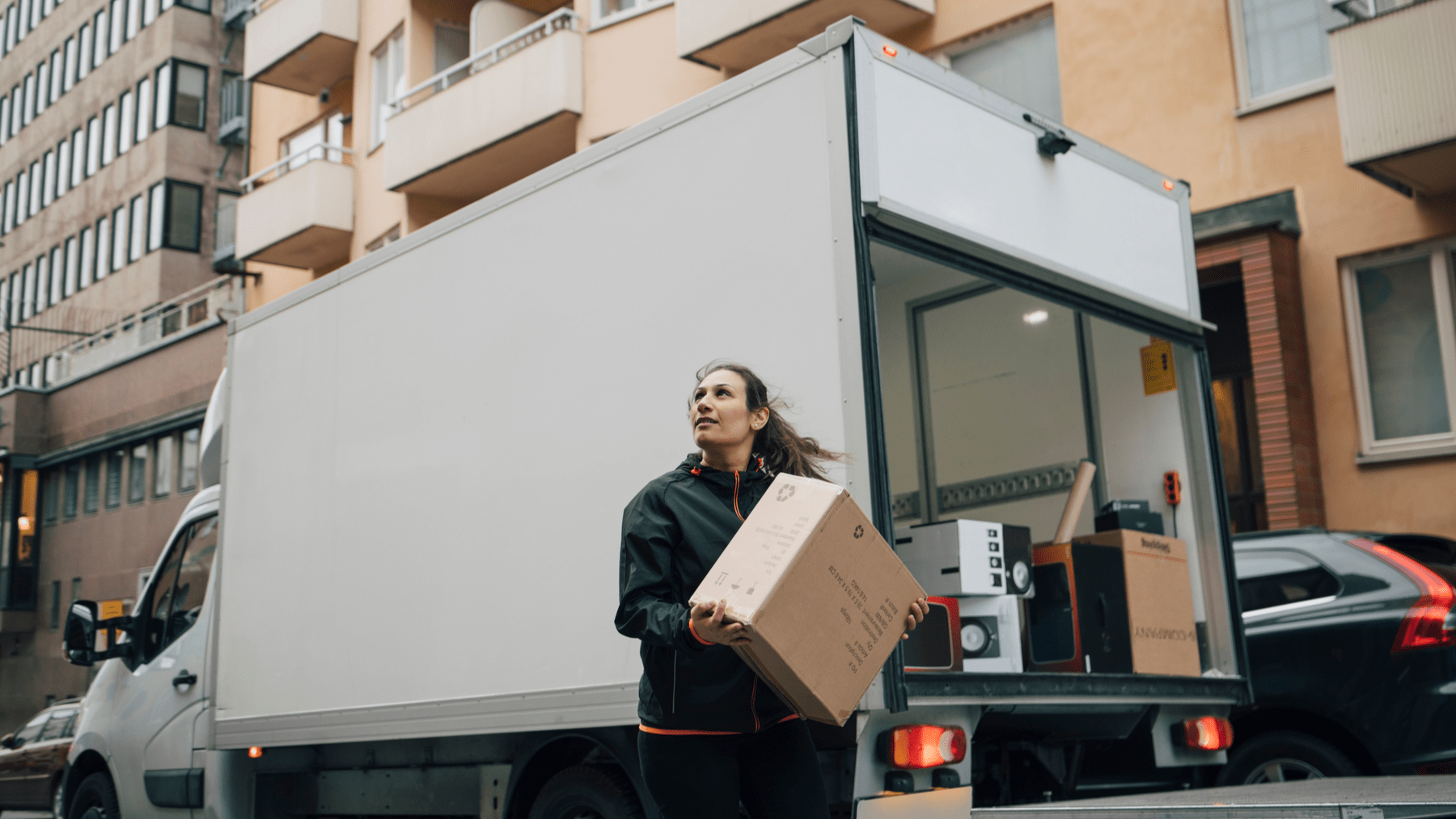Person carrying a cardboard box from the back of a white delivery truck parked on a city street. Apartment buildings and parked cars are visible in the background, and additional moving boxes and household items are loaded inside the truck.
