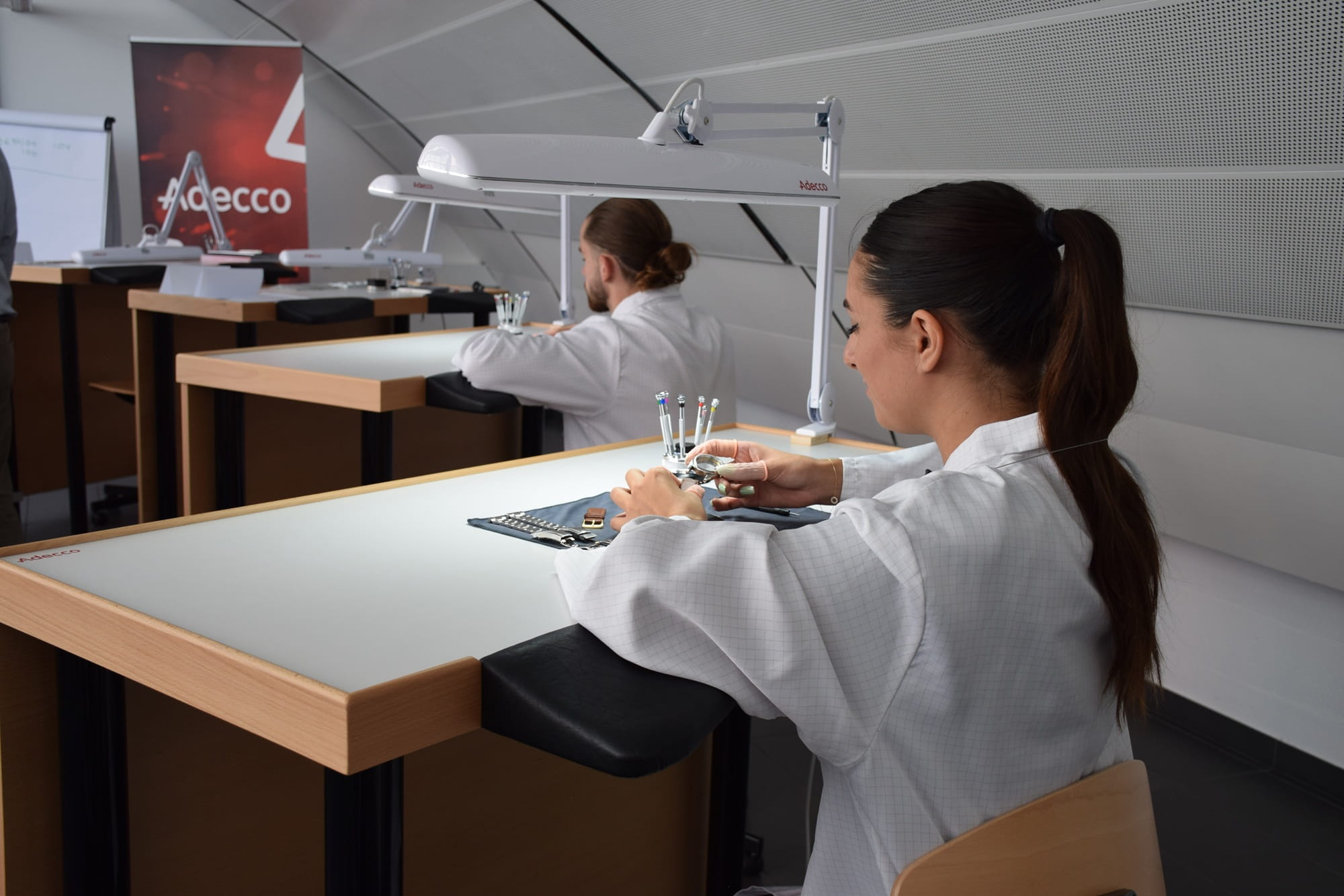 Studentinnen einer Uhrmacherschule beim praktischen Lernen mit Uhrwerk-Komponenten und Werkzeugen in einem Ausbildungsraum. Studentinnen einer Uhrmacherschule beim praktischen Lernen mit Uhrwerk-Komponenten und Werkzeugen in einem Ausbildungsraum.
