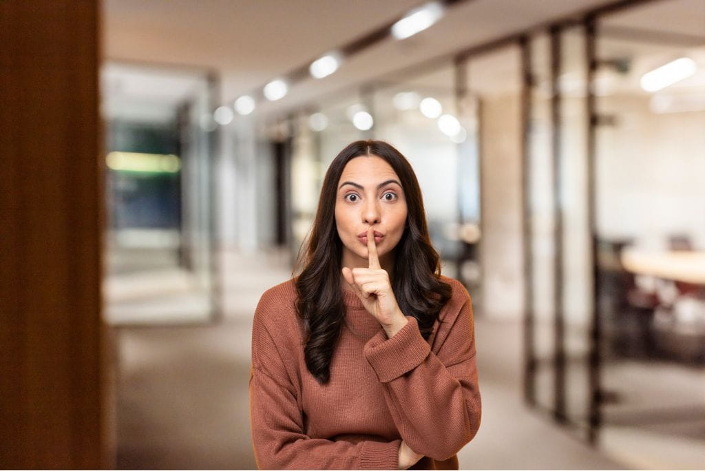 Eine Frau im Büro hält den Zeigefinger an die Lippen und macht eine „Psst“-Geste.