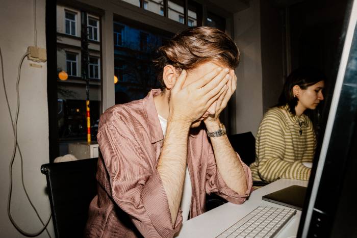 Person sitzt an einem Schreibtisch vor einem Computer und hält erschöpft die Hände vors Gesicht.