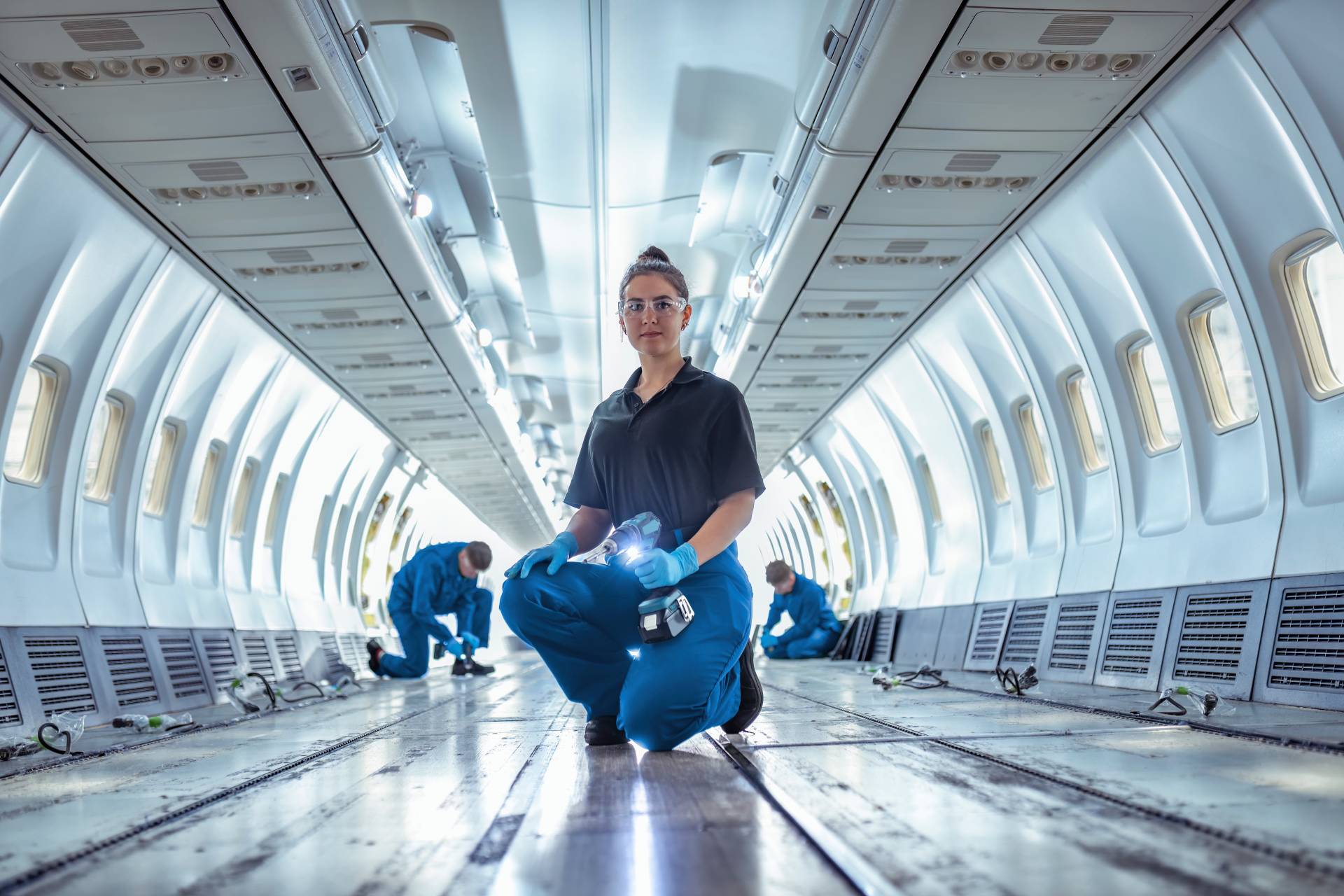 Eine Flugzeugtechnikerin kniet im Innenraum eines Flugzeugs und hält einen Akkuschrauber. Die Sitze sind ausgebaut, und Wartungsarbeiten werden im hell beleuchteten Flugzeugrumpf durchgeführt.