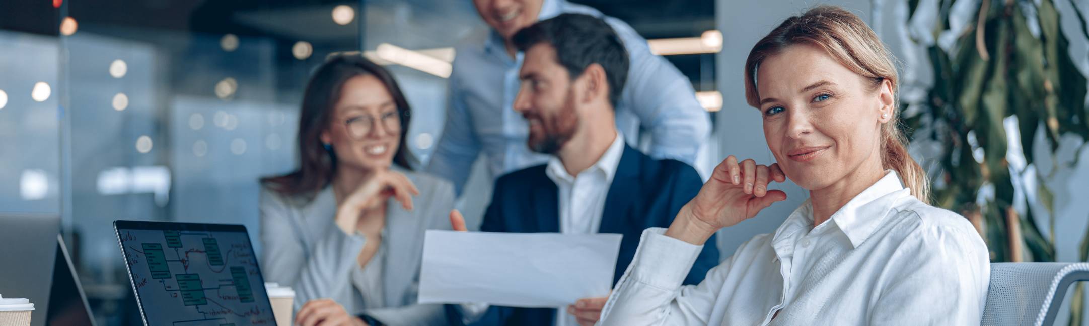 Teammeeting in einem modernen Büro mit vier Geschäftspersonen; eine Frau im Vordergrund schaut in die Kamera, während im Hintergrund Kolleg:innen zusammenarbeiten.