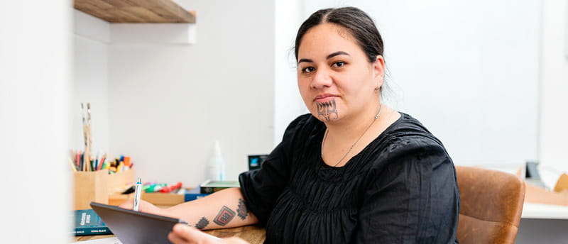 Person with Māori tāmoko tattoos working at a desk with a pen and tablet in an office setting