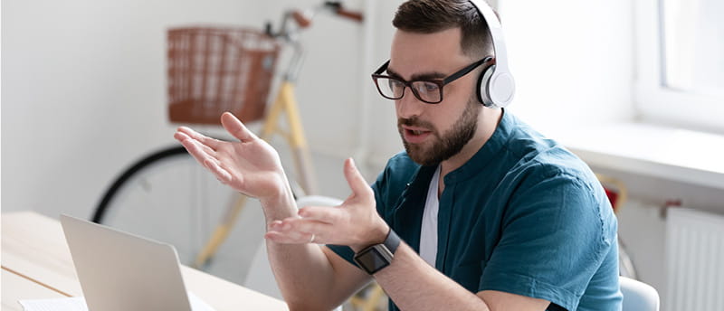 Person on a video call at home, wearing headphones and gesturing while seated at a desk with a laptop