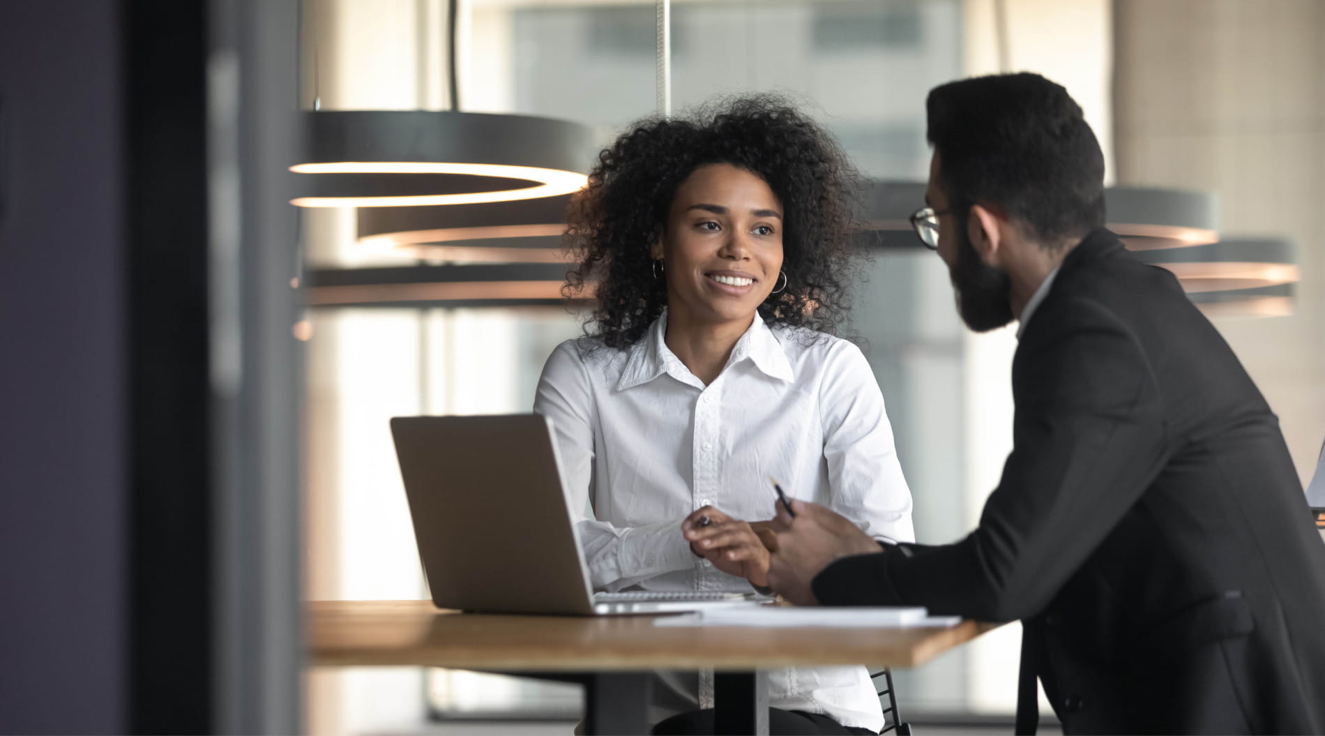 Two people in professional attire having a discussion at a table with a laptop in an office setting