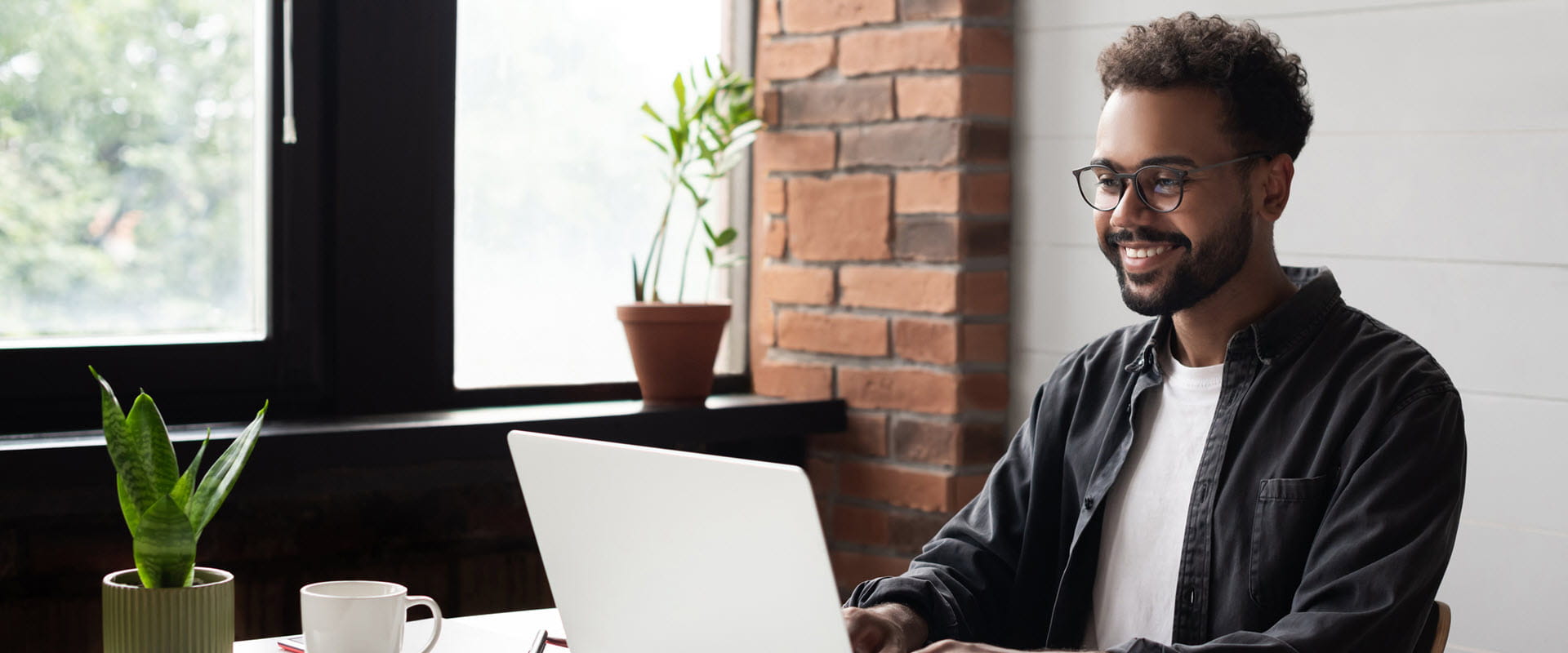 Smiling person wearing glasses sits at a desk with a laptop.