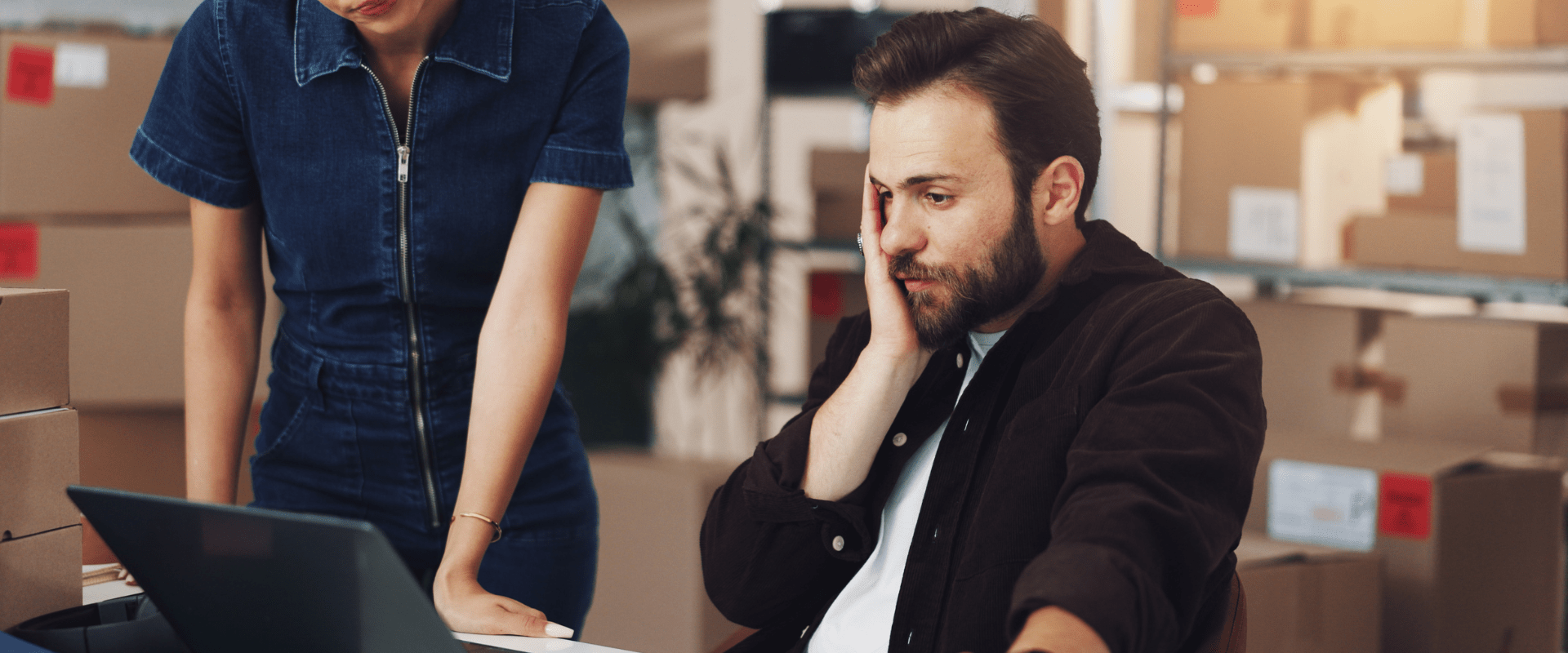 A person sits at their desk looking at a laptop, while someone stands next to them.