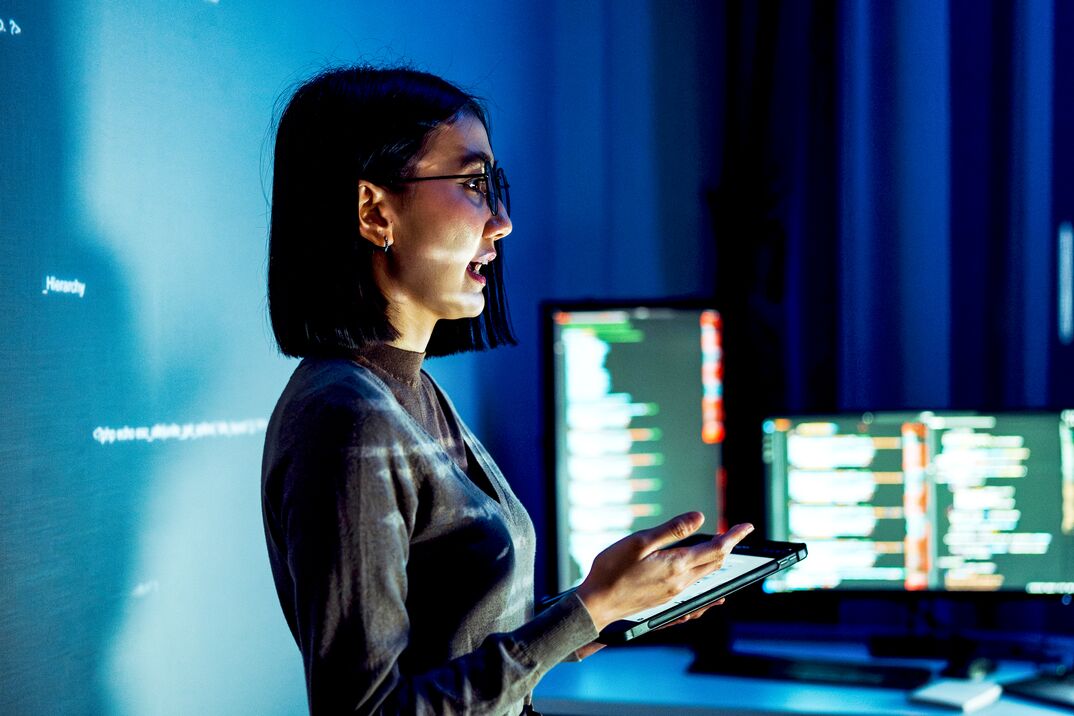 Person holding a tablet in a dim office with multiple monitors displaying code. Person holding a tablet in a dim office with multiple monitors displaying code.