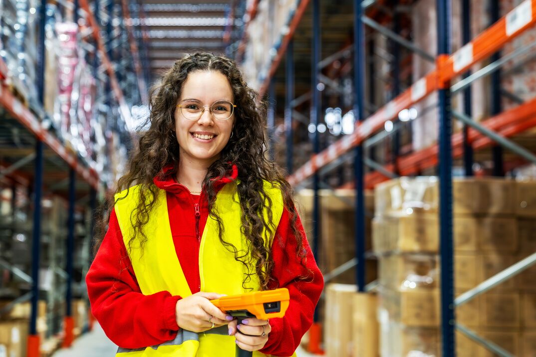 Person wearing a high-visibility safety vest and red jacket holding a handheld scanner while standing in a warehouse Person wearing a high-visibility safety vest and red jacket holding a handheld scanner while standing in a warehouse