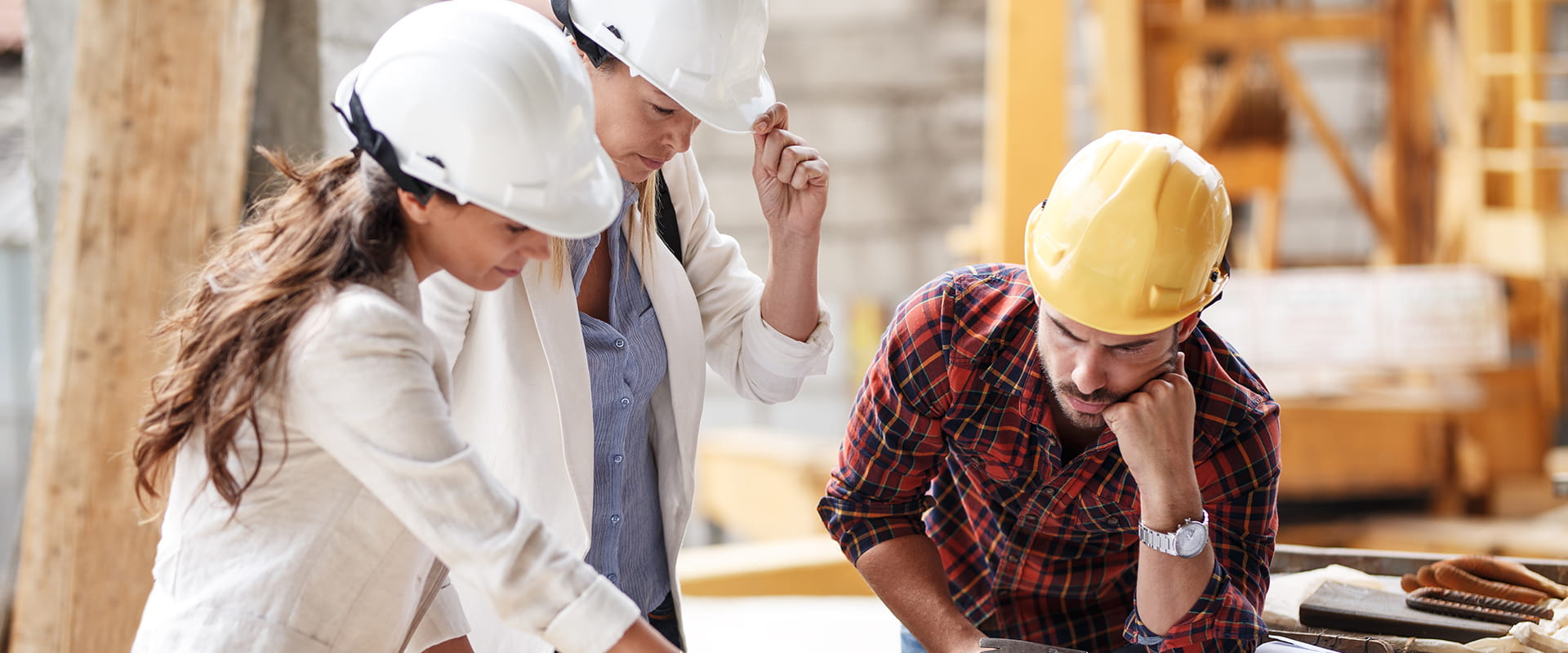 Three construction workers in hard hats examine plans on a construction site, focused on their discussion and tasks.
