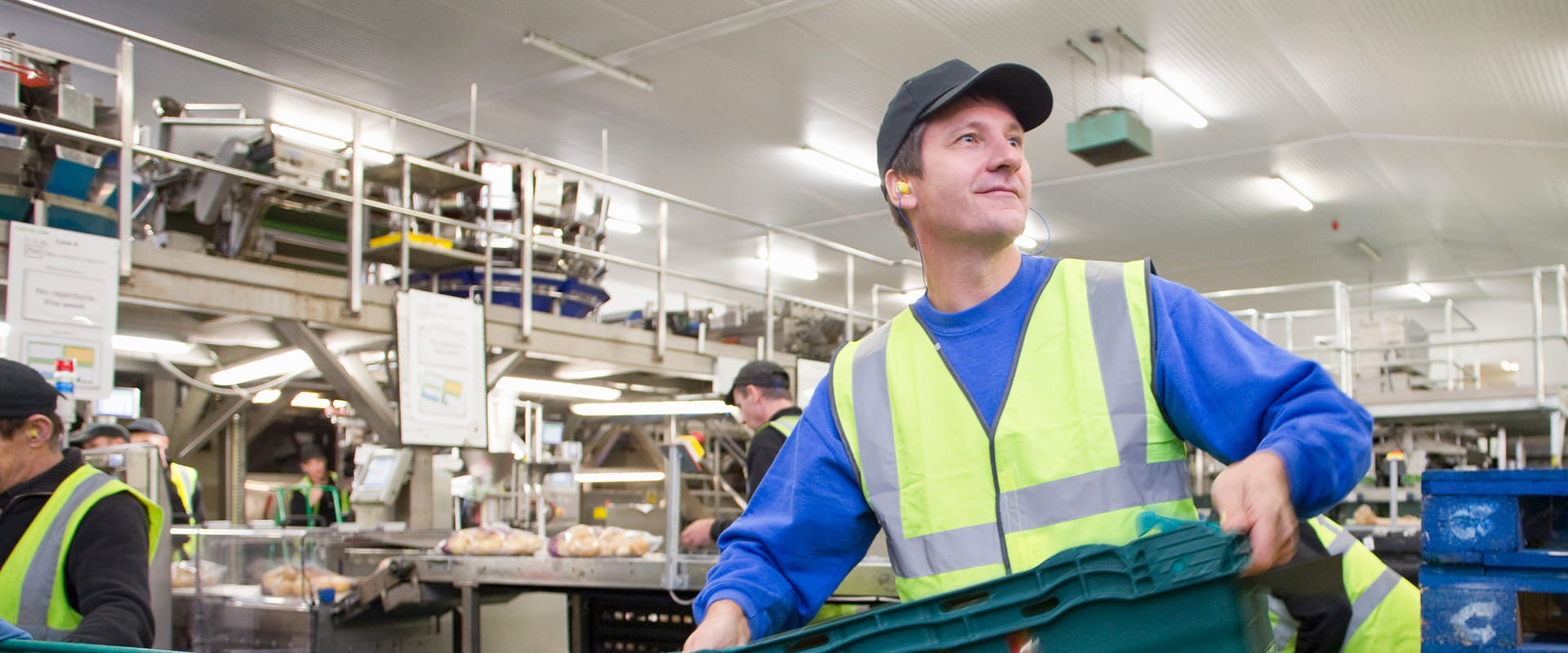 A warehouse worker in a safety vest and cap handles crates in a busy industrial setting with machinery and other workers.
