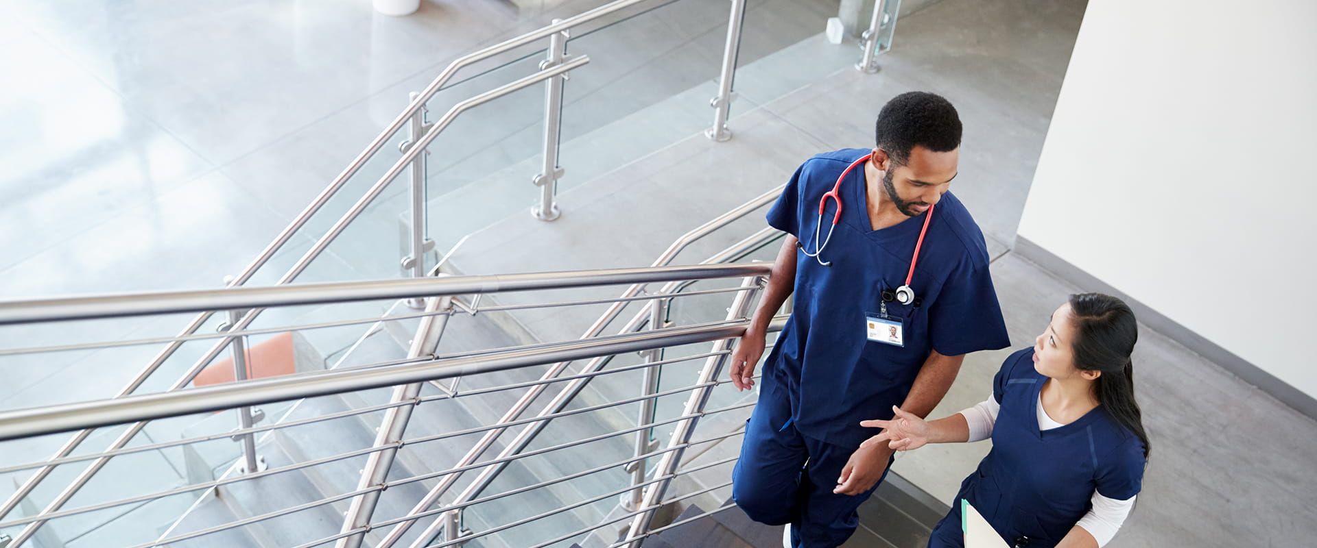 Two healthcare professionals in scrubs walk and talk up a staircase in a modern medical facility, discussing their work.