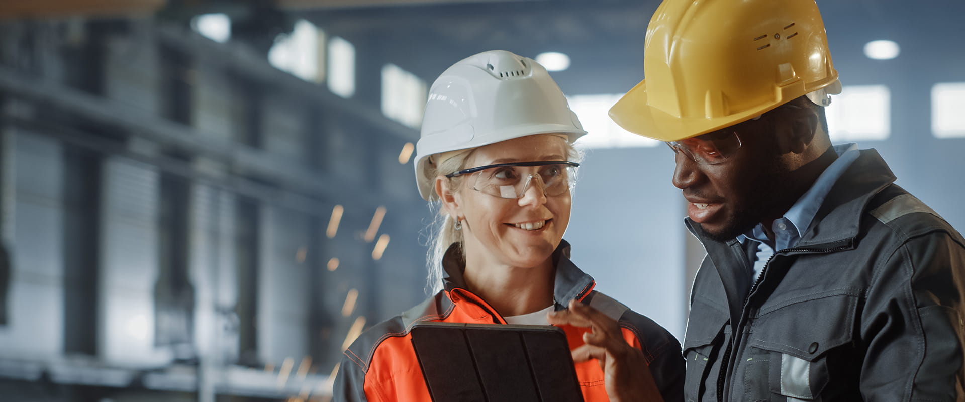 Two engineers in hard hats and safety gear discuss a project while looking at a tablet in an industrial facility.