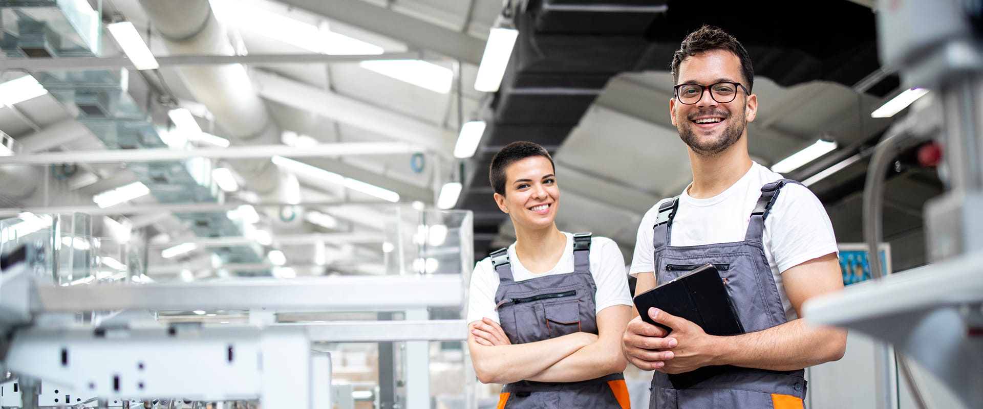 Two smiling workers in overalls stand in a modern factory, one holding a tablet. They appear confident and professional.