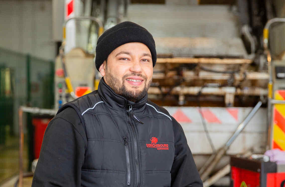 Smiling worker in a black vest and beanie standing in front of a garbage truck, ready for duty, in an industrial environment with tools and equipment visible
