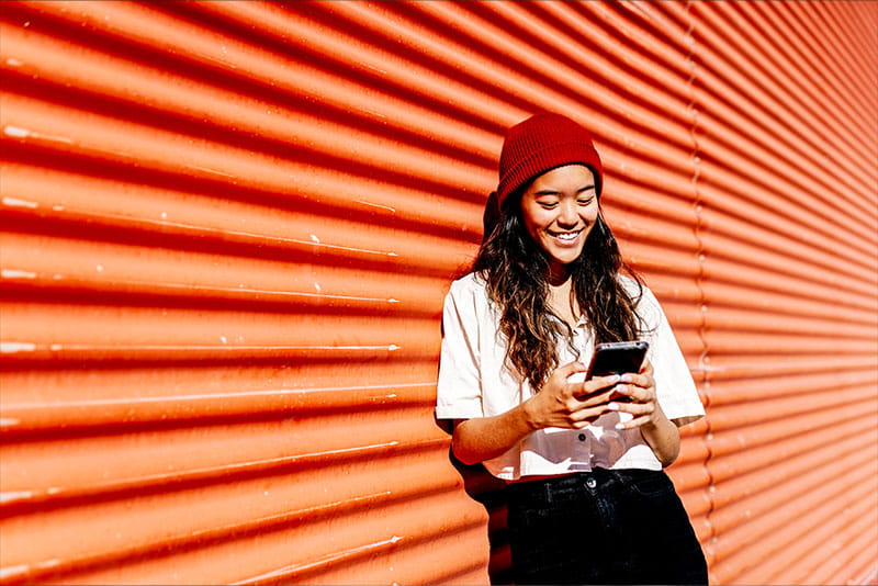 Young woman in a red beanie smiling while using her smartphone, standing against a bright orange corrugated wall.