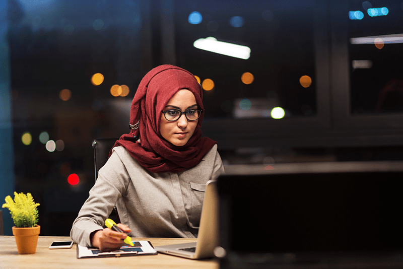 Job seeker working on a laptop in a modern office at night, taking notes while researching career opportunities and preparing applications online.