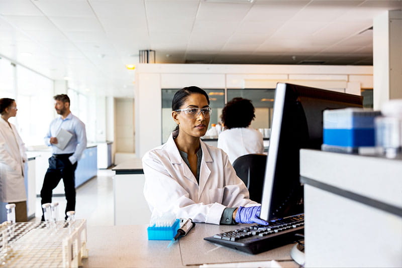 Scientist working at a computer in a modern laboratory while colleagues collaborate in the background.