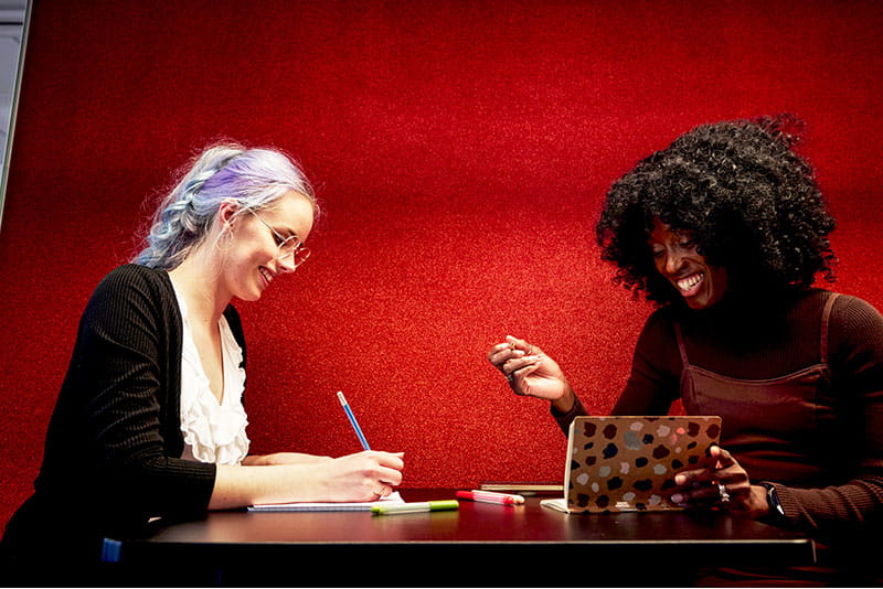 Two colleagues collaborating at a table, one writing notes while the other uses a laptop, against a bright red background.