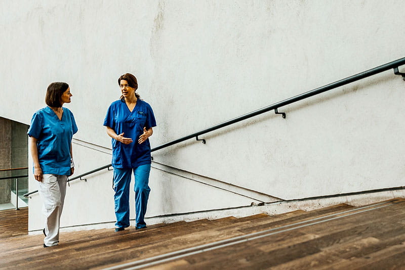 Two healthcare professionals in scrubs walking up stairs, talking and collaborating in a clinical setting.