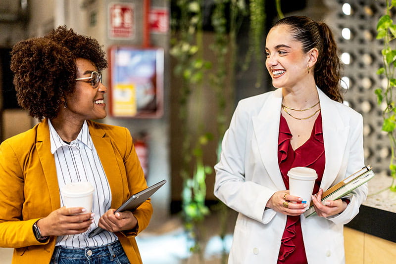 Two professionals walking through a modern office, smiling and chatting while holding coffee cups and work materials.