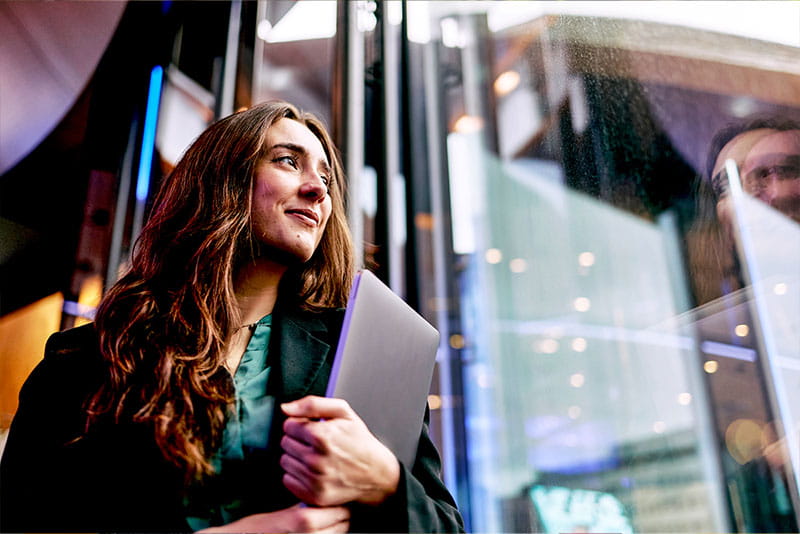 Professional holding a laptop while standing by a glass window, looking outside in a modern office building. Professional holding a laptop while standing by a glass window, looking outside in a modern office building.