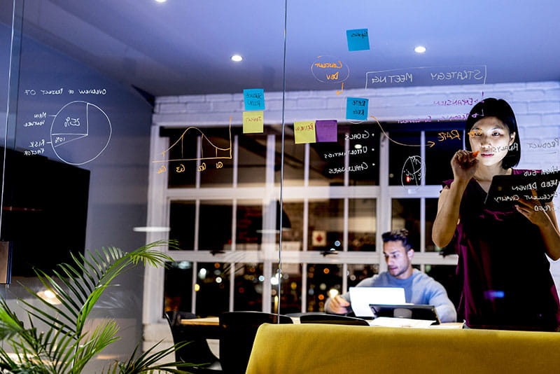 Professional writing ideas on a glass board during a planning session while a colleague works in the background of a modern office.