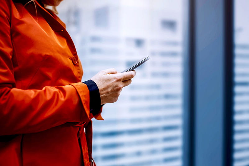 Person in a red coat using a smartphone while standing outdoors in an urban setting with blurred buildings in the background.