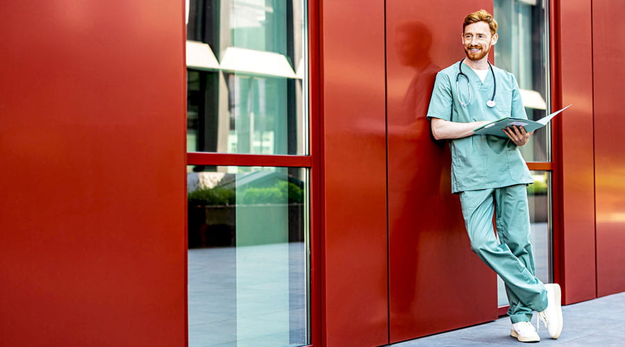 Soignant en tenue médicale appuyé contre un mur rouge avec un dossier à la main