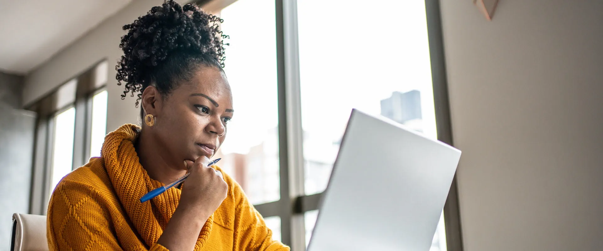 Focused professional working on a laptop at home, wearing a yellow sweater, holding a pen Focused professional working on a laptop at home, wearing a yellow sweater, holding a pen