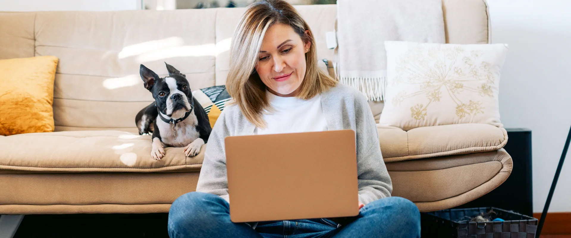 A person sits on the floor, leaning against a couch, with their laptop in their lap. A dog sits behind them on the couch.