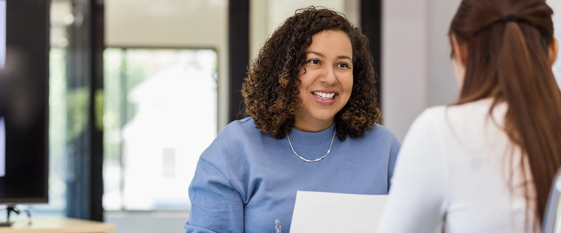 A cheerful woman with curly hair talking to a colleague in a modern office setting, holding a document.