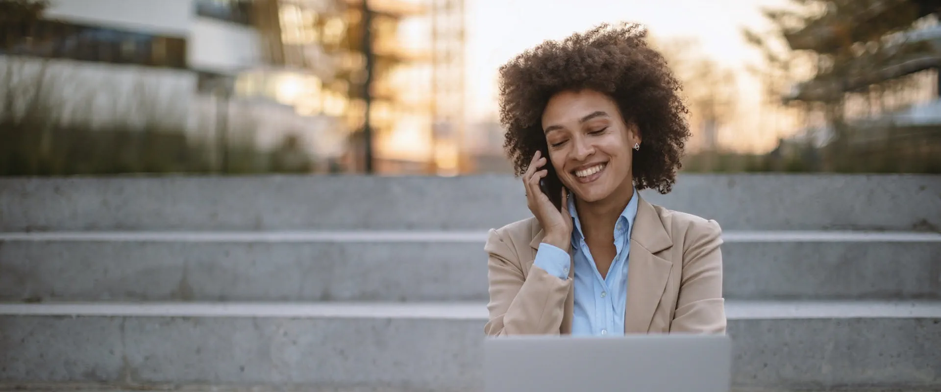 Person sitting outdoors on steps, engaged in a phone conversation while working on a laptop, smiling in a professional setting. Person sitting outdoors on steps, engaged in a phone conversation while working on a laptop, smiling in a professional setting.