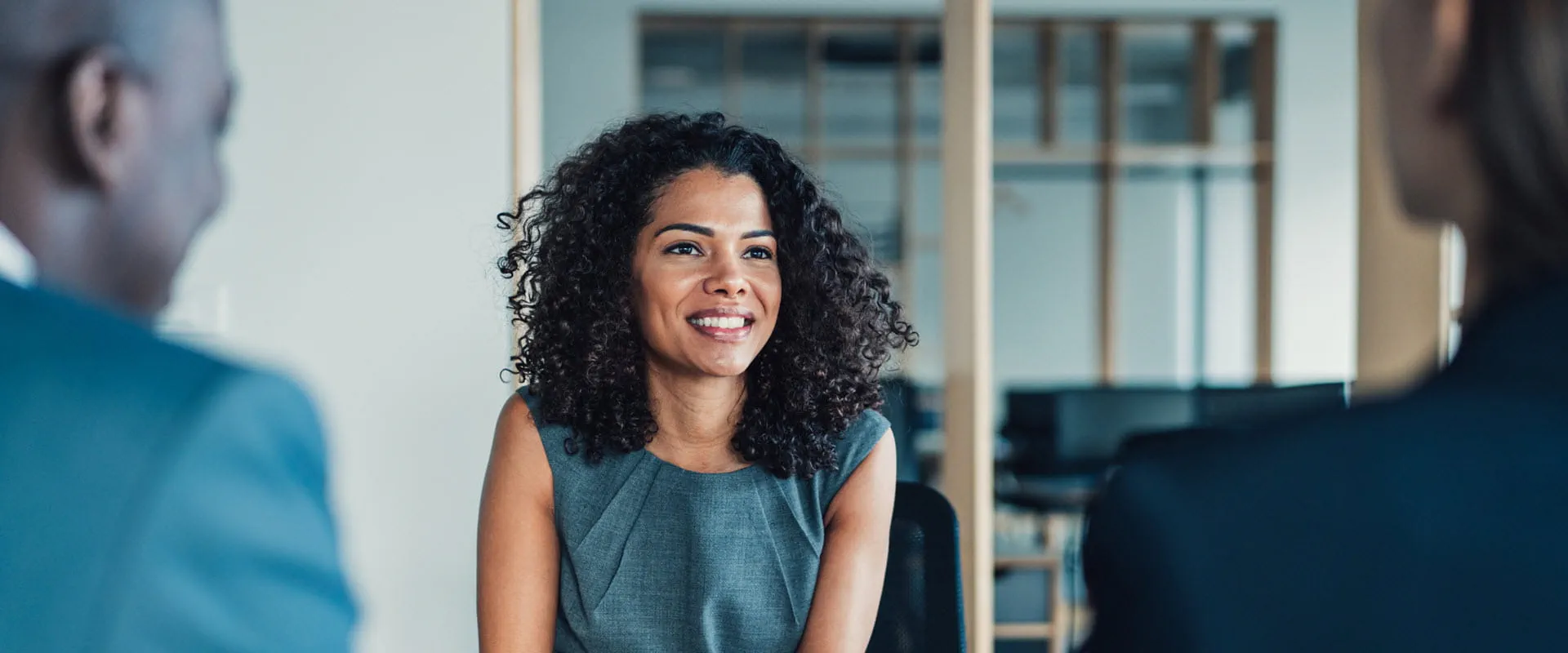 A confident professional sitting in a meeting room, engaging with colleagues who are partially visible. A confident professional sitting in a meeting room, engaging with colleagues who are partially visible.