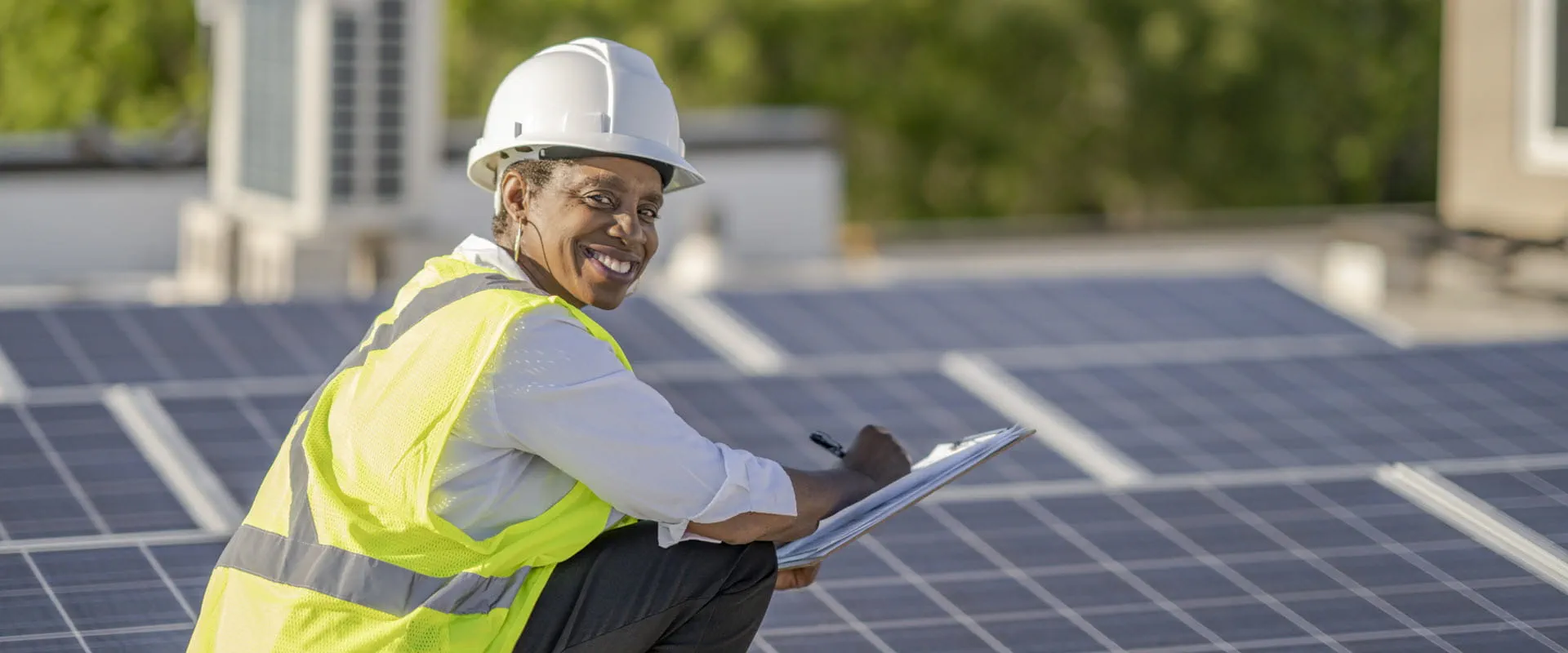 A person in a hard hat and high-visibility vest smiles while inspecting solar panels on a rooftop, holding a clipboard and pen.