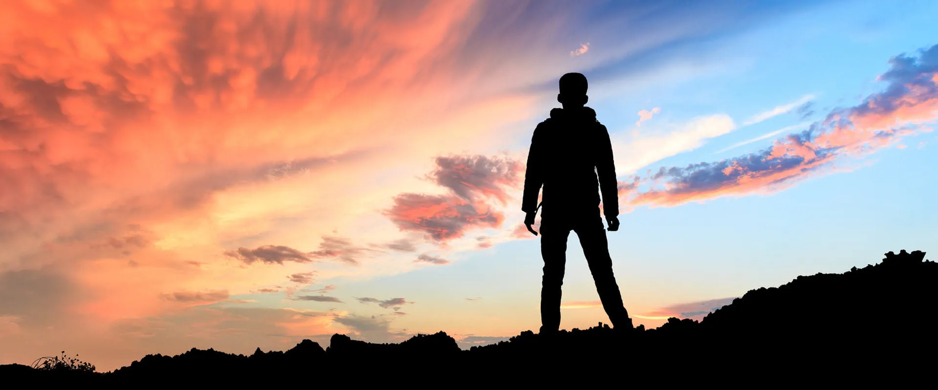 Silhouette of a person standing on a hilltop against a dramatic sunset sky with vibrant orange, pink, and blue hues. Silhouette of a person standing on a hilltop against a dramatic sunset sky with vibrant orange, pink, and blue hues.