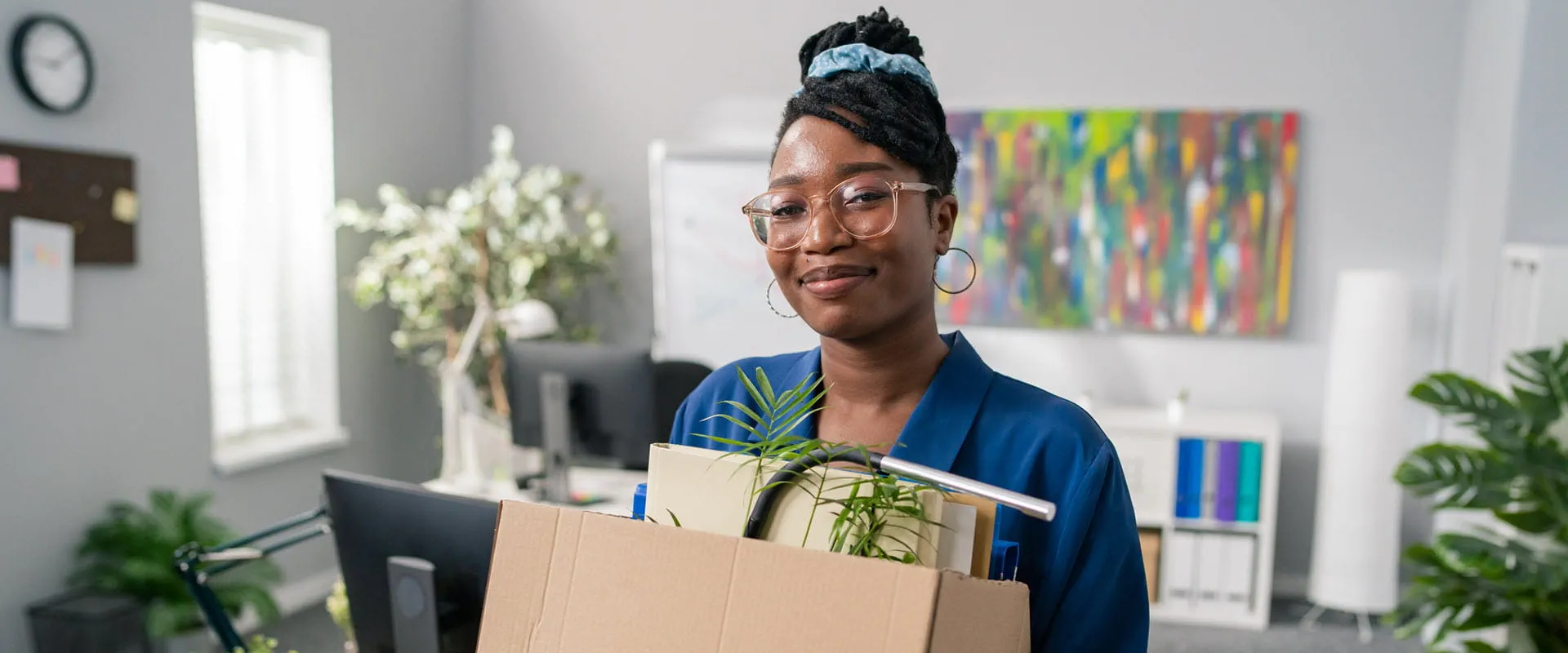 After quitting, a smiling individual walks out of an office with a box of files and plant After quitting, a smiling individual walks out of an office with a box of files and plant