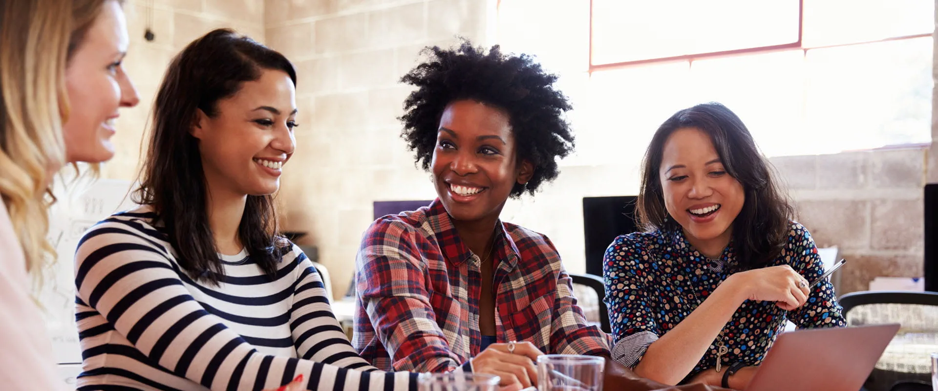 Four people sitting at a table, smiling and collaborating while using a laptop in a casual office setting. Four people sitting at a table, smiling and collaborating while using a laptop in a casual office setting.