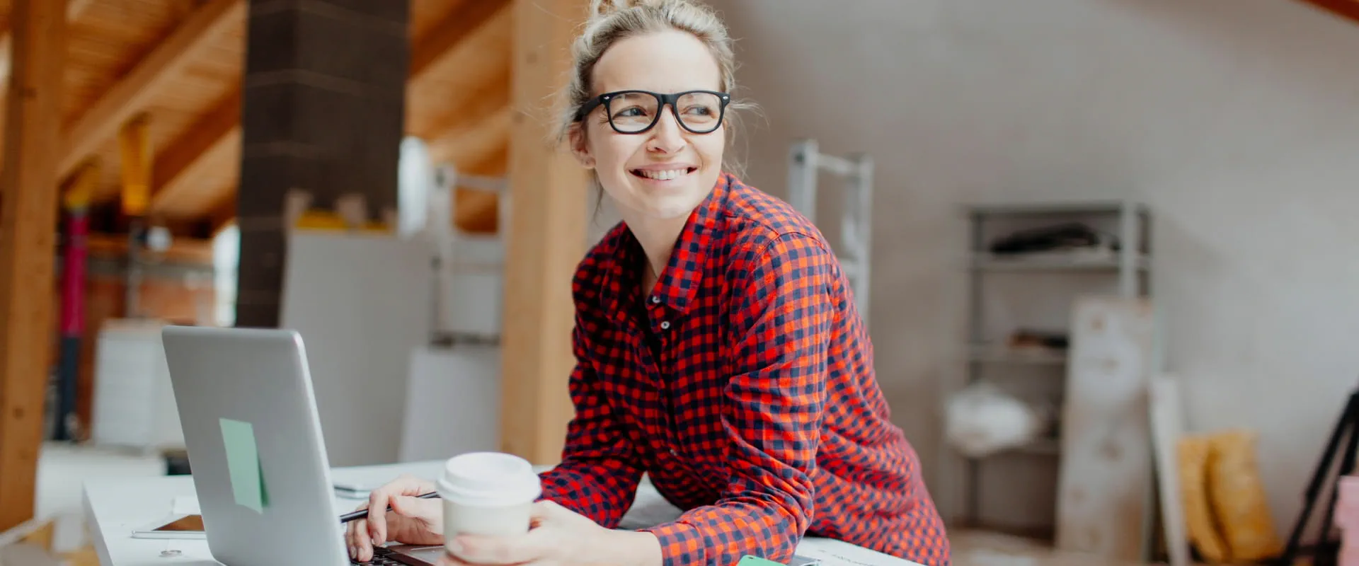 A creative professional working on a laptop in a stylish loft office setting, holding a coffee cup.