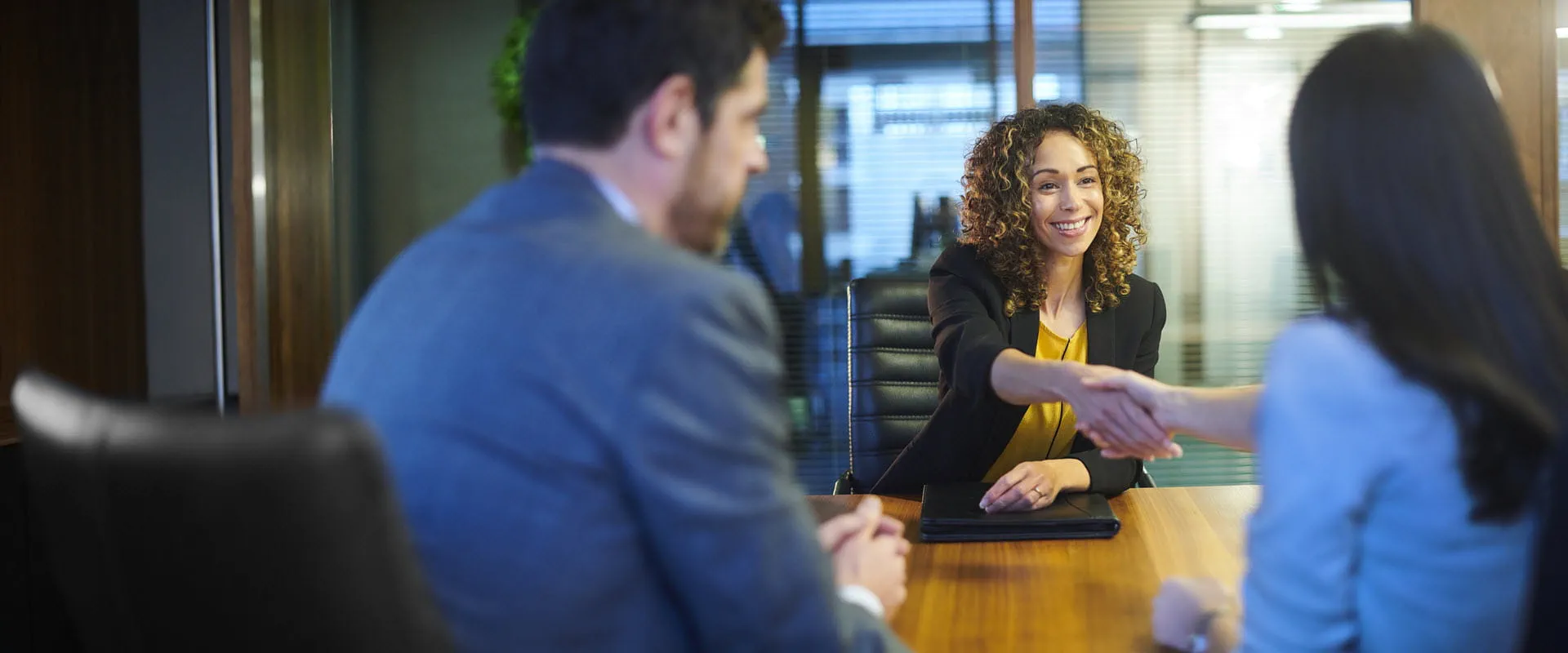 A smiling individual shakes hands across a boardroom table with one of their two interviewers.