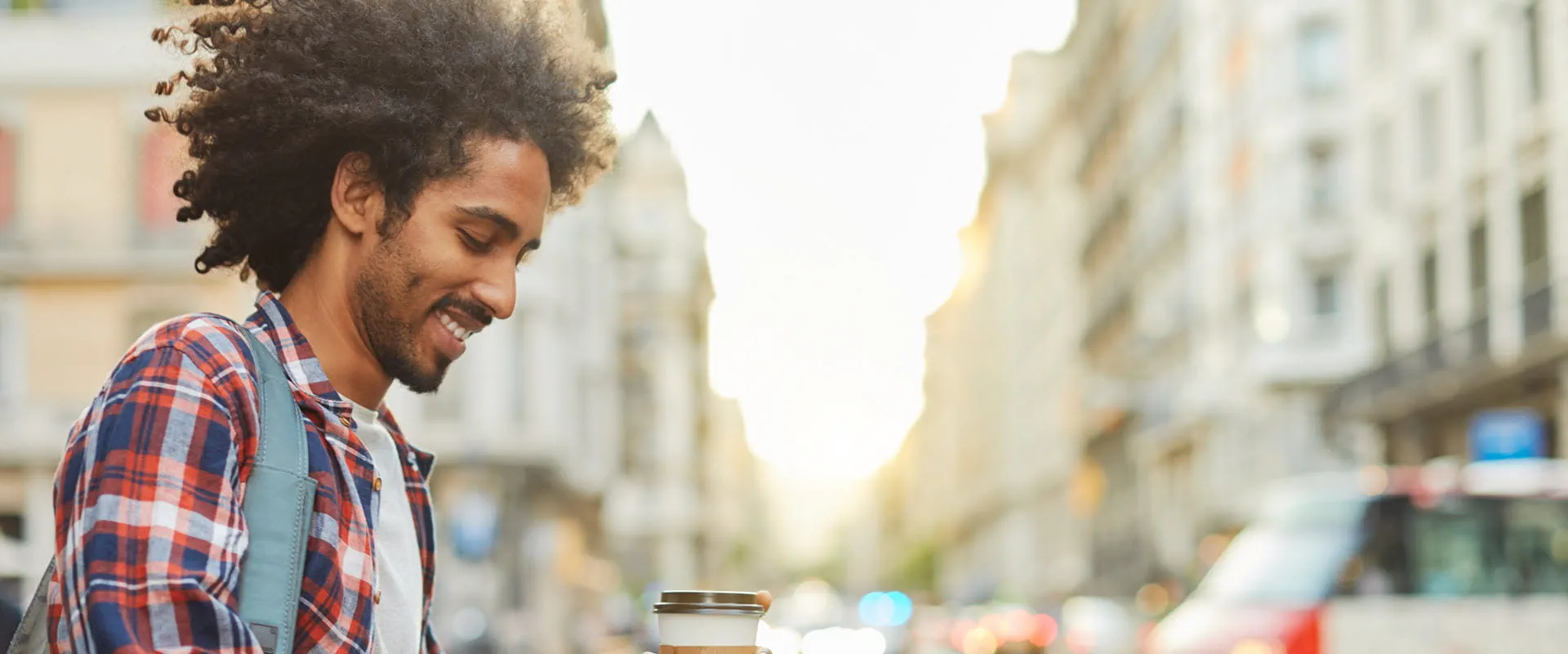 A smiling individual walks down a city street, looking at his phone and holding a cup of coffee. A smiling individual walks down a city street, looking at his phone and holding a cup of coffee.