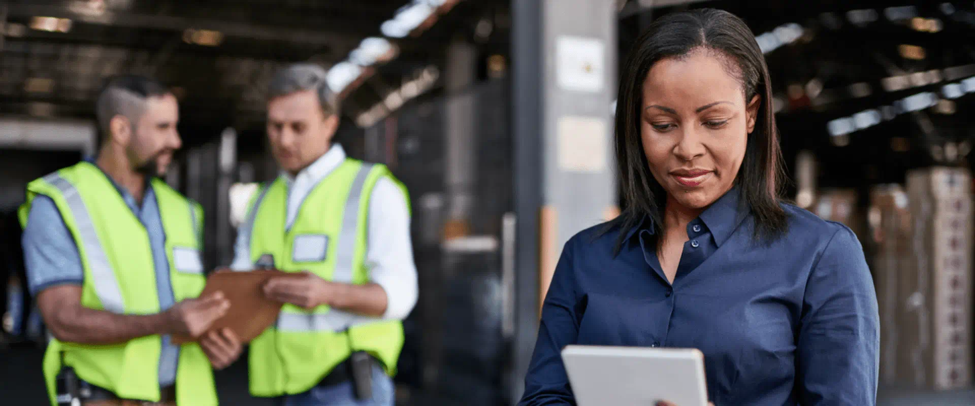 A worker looks at a tablet, while two others in safety vests talk behind them. A worker looks at a tablet, while two others in safety vests talk behind them.