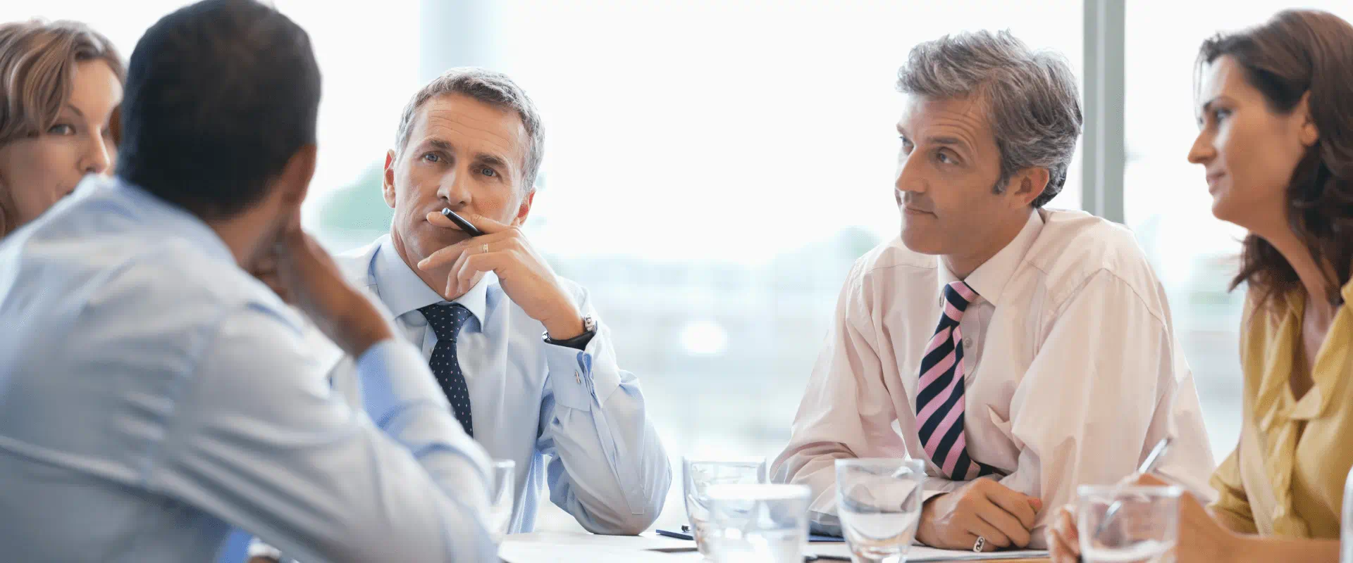 Five business people sit at a conference table listening to someone talk. 
